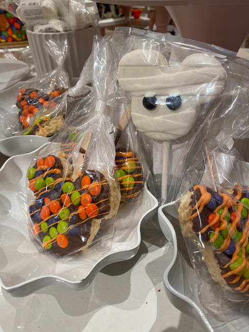 Colorful chocolate treats wrapped in plastic, featuring Mickey-shaped marshmallow pops and candy-coated rice treats, displayed on a wavy white dish.