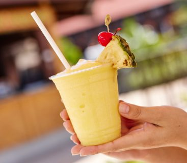 A hand holds a yellow smoothie with a straw, garnished with pineapple and cherry. Blurred background with warm colors suggests a tropical vibe.