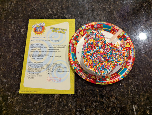 Colorful Mickey-shaped treat with sprinkles on a festive plate next to a "Create Your Own Treat" menu on a dark countertop.