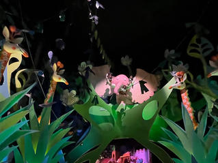 Colorful animated jungle scene with giraffes and elephant surrounded by large green leaves and flowers. Playful, whimsical atmosphere.