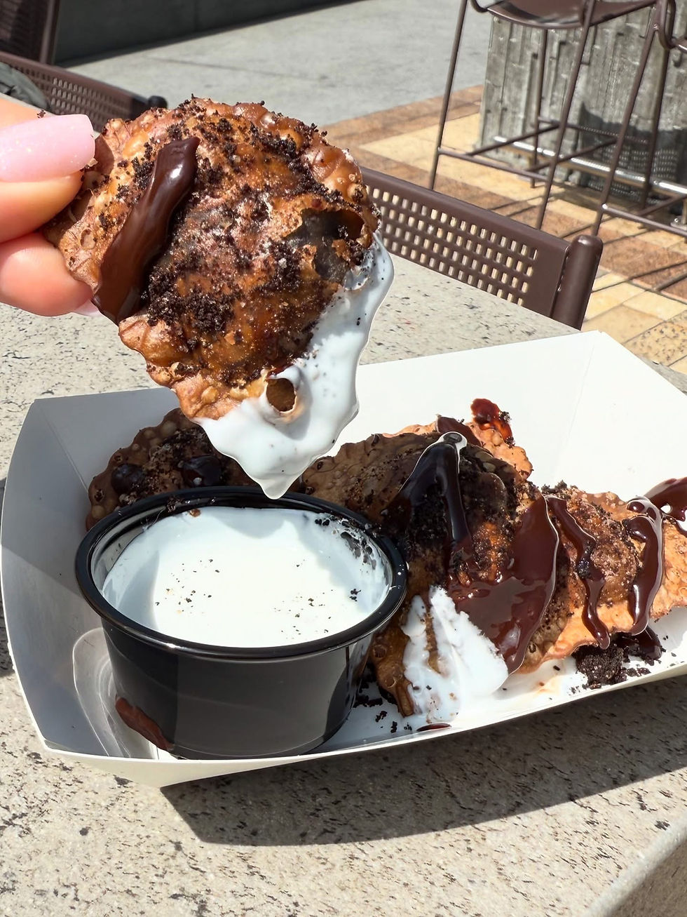 A hand dips a chocolate-drizzled fried cookie into white sauce. Sunlit outdoor table with metal chairs in the background.
