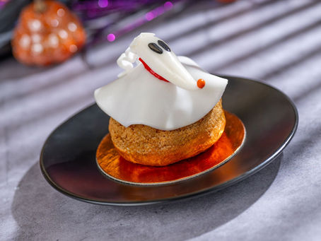Halloween-themed pastry with a white ghost decoration sits on a dark plate. Blurred orange pumpkin and purple highlights in the background.