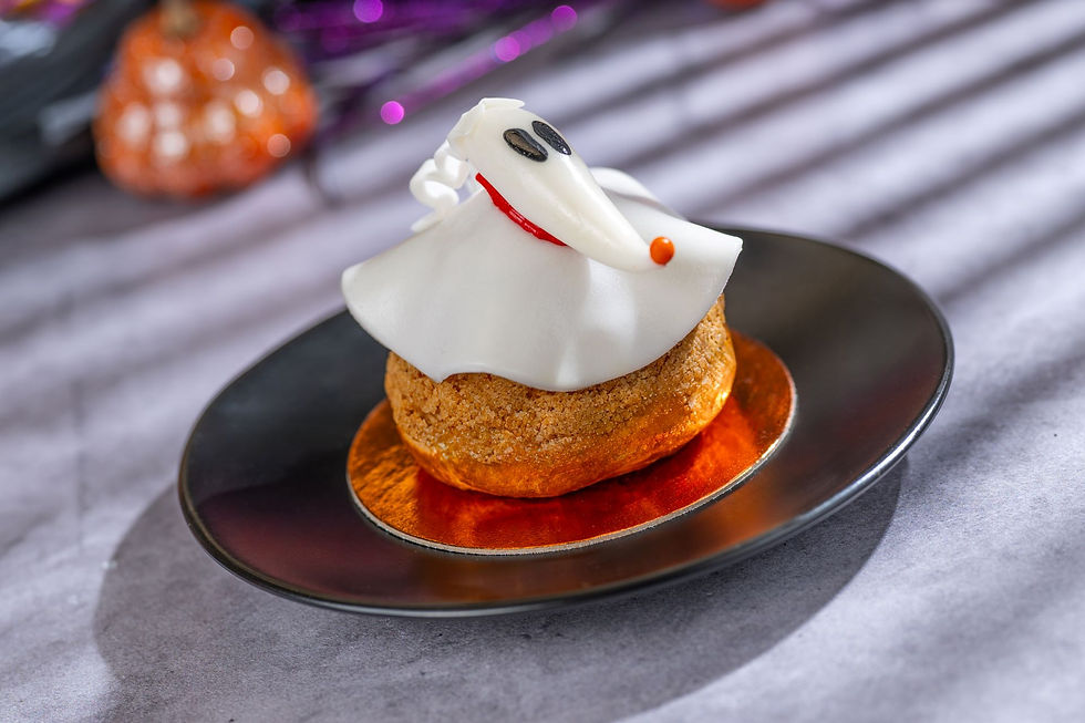 Halloween-themed pastry with a white ghost decoration sits on a dark plate. Blurred orange pumpkin and purple highlights in the background.