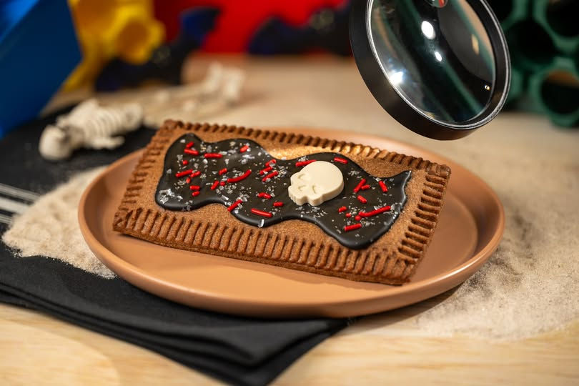 Dessert on a tan plate with chocolate icing, sprinkles, and a white skull decoration. Background has a skeleton toy and colorful objects. Spooky vibe.