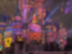 Colorful castle with projections, blue turrets, and laser lights. Jack-o'-lantern in front, night sky with fireworks. Festive atmosphere.