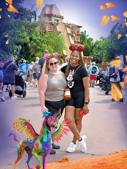 Two women wearing Disney-themed attire smile at a theme park. A colorful, animated creature stands beside them. Bright, festive atmosphere.