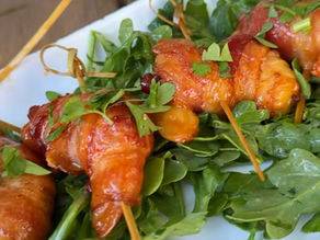 Skewered bacon-wrapped apricots on fresh greens, garnished with herbs, served on a white plate. Bright and appetizing presentation.