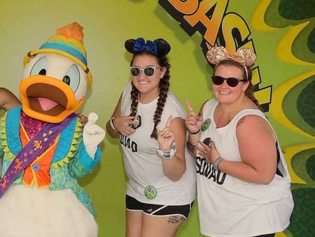 Two women in white "Squad" shirts pose with a colorful duck character. They're smiling with finger gun gestures. Green "Dino-Bash" backdrop.