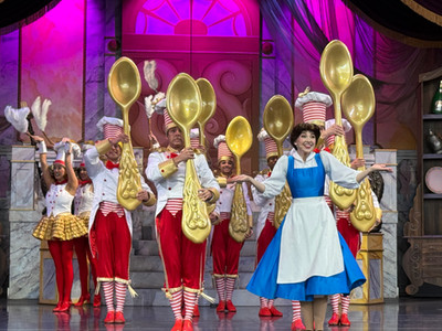 Performers in colorful costumes, including a woman in a blue apron, dance holding large golden spoons on a vibrant stage with pink lighting.