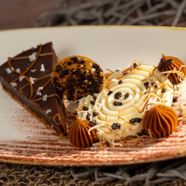Chocolate tart with sea salt next to white frosting spiral, caramel dollops, and chocolate garnish on a white plate. The creamy and elegant Brigadeiro Prestígio at Jungle Navigation Co. LTD Skipper Canteen, at Disney's Magic Kingdom.