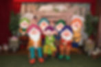 Seven dwarfs in colorful outfits pose with two people, smiling in front of a Mine backdrop. Gem barrels and green foliage frame the scene.
