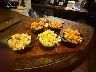 Assorted popcorn in black bowls on a wooden tray at a bar counter. A drink and menu are nearby. Warm lighting creates a cozy mood.