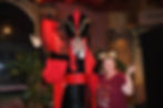 A woman in Mickey ears poses with a costumed character in a red and black outfit. Background includes plants and a brick wall. Playful mood.