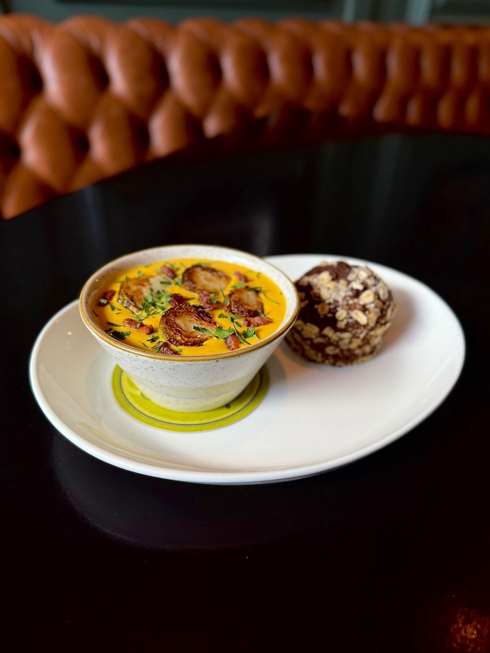 Creamy soup with herbs and crispy toppings in a bowl on a white plate, next to a seeded bread roll. Background: brown cushioned seat.