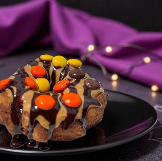 Donut with chocolate and colorful candies on a black plate. Purple fabric and warm fairy lights in the background create a cozy mood.