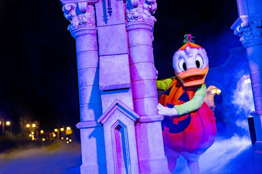 Cartoon duck in a pumpkin costume peeks from behind a stone pillar. Blue and purple lighting with fog creates a spooky atmosphere.