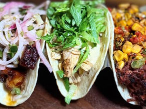 Three tacos on a wooden board: one with pickled onions, one with lettuce and chicken, and one with corn and peppers, vibrant and appetizing.