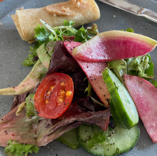 A fresh salad with greens, tomato, radish, and cucumber on a gray plate is next to a rolled bread piece. A knife rests beside the bread.