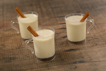 Three clear mugs of creamy eggnog with cinnamon sticks on a rustic wooden table. Specks of spice are visible, creating a cozy feel.
