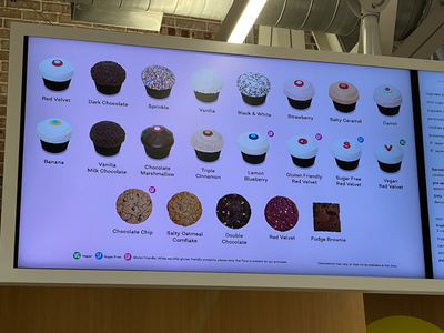 Two frosted cupcakes in a Sprinkles box on a white surface. One has a red "G" decoration, the other a black and blue circle. Mood: indulgent.