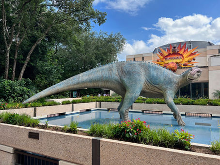 A dinosaur statue stands in a garden near a building with a "DINOSAUR" sign and explosion art. Green trees and a bright sky are in the background.