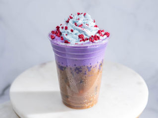 Colorful drink with a brown and purple gradient in a cup, topped with blue whipped cream and red sprinkles. Set on a white surface.