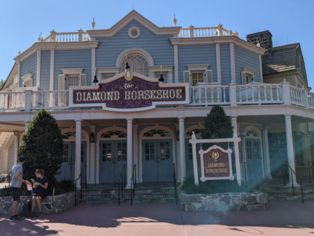 Two people sit and stand near The Diamond Horseshoe building. Blue sky, ornate facade, brown sign, and greenery enhance the calm setting.