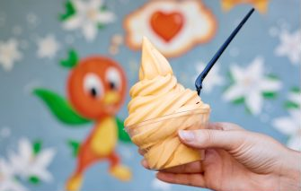 Hand holds orange swirl ice cream with black spoon. Background has cartoon orange character and colorful patterns, creating a playful mood.