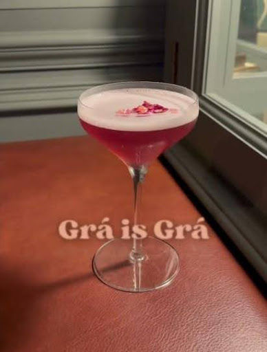 A pink cocktail with a frothy top and flower petals sits on a brown leather surface. Text reads “Grá is Grá.” The mood is elegant. This is the Raglan Road Pride Cocktail at Disney Springs.