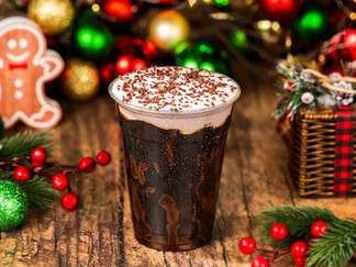Festive scene with iced coffee topped with cream and chocolate sprinkles, surrounded by Christmas ornaments and a smiling gingerbread.