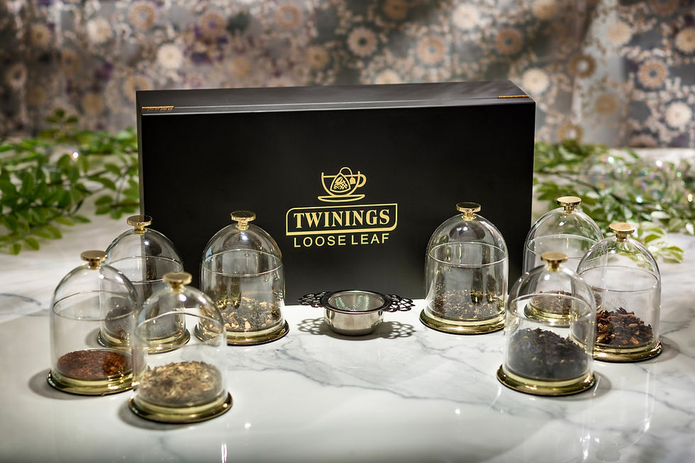 Tea display with eight glass domes containing loose leaves, centered around a black "Twinings Loose Leaf" box. Floral backdrop.