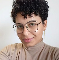 a imagem mostra uma mulher negra de pele clara com cabelos cacheados e curtos e olhos verdes. Ela veste uma blusa bege com gola, e óculos quadrados.