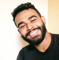 imagem mostra um homem negro de pele clara, com cabelos cacheados e curtos, barba cheia e sorriso aberto. Ele veste uma camiseta preta.