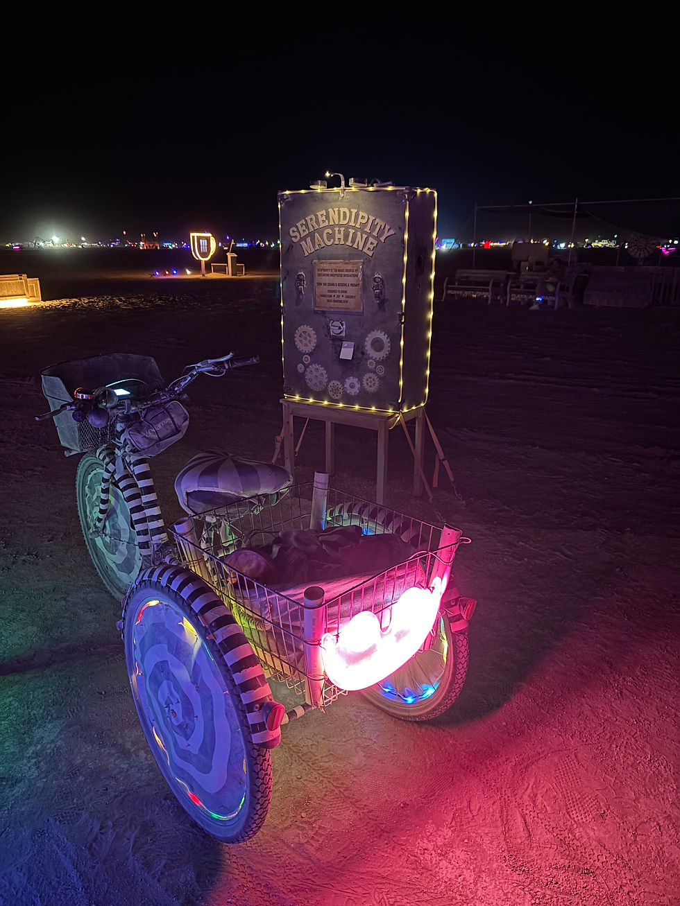 Tricycle with colorful lights and a "Serendipity Machine" sign in a dimly lit desert setting, with scattered distant lights.