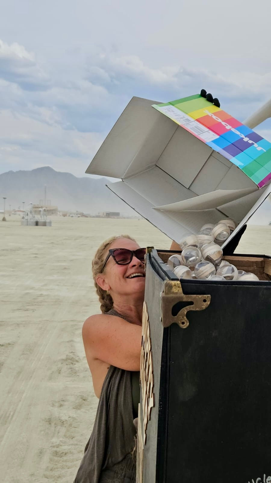 A smiling person pours clear spheres from a colorful box into a black container which is a Serendipity Machine in a dusty, open desert Burning Man setting with mountains in the background.