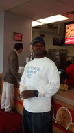 Man stands in a restaurant, wearing a hat and a white shirt with text.