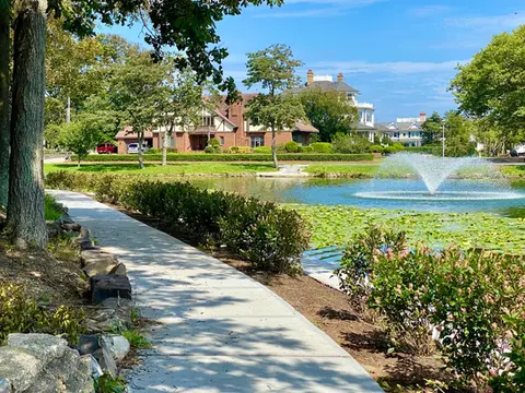 Pathway alongside a pond with fountain, trees, and distant houses. | Down To Earth Landscaping | Jackson New Jersey, USA (732) 833-7702