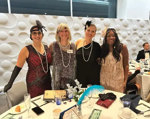 Four women in 1920s flapper dresses smiling at an event.