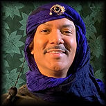 Brown skin, smiling man wears blue turban with black tassel on the right and golden-eye opal and pearl stick pin centered.