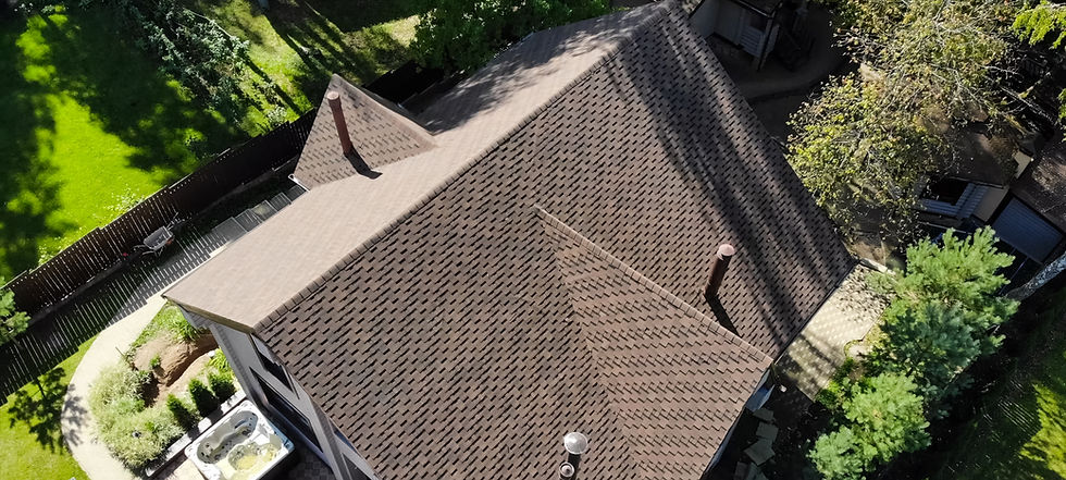Aerial view of a house with a brown patterned roof, surrounded by trees and a green lawn. A backyard hot tub is visible. Sunny day.
