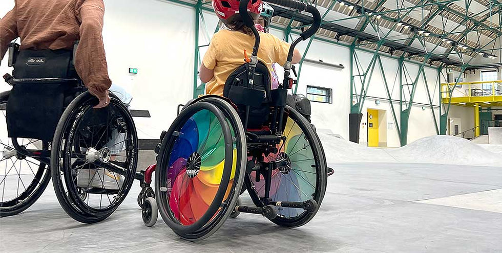Ein bunter Rollstuhl mit tollen Regenbogenfarben an den Rädern. Das Mädchen blickt auf den Skatepark.