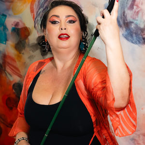 Clarice Senna, mulher branca de cabelos lisos escuros, segura bengala diante de pintura colorida, vestindo roupa laranja e preta.