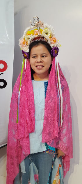 Sabrina Santos sorrindo usa coroa de flores e véu rosa figurino de uma apresentação sua em peça teatral.