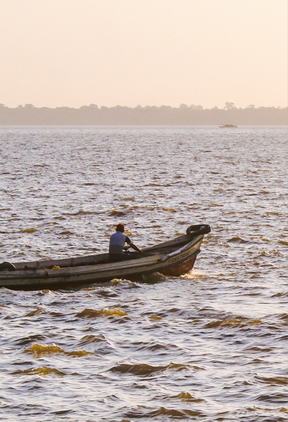 Homem navegando de barco em rio; céu ensolarado e vegetação ao fundo. Fotografia de Matheus Sousa.