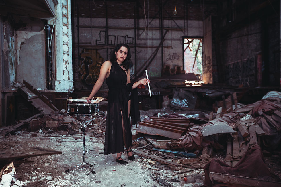 A female drummer juggler in a sheer, black dance dress standing beside a snare drum with drumsticks in a dilapidated, graffiti-ridden auditorium.