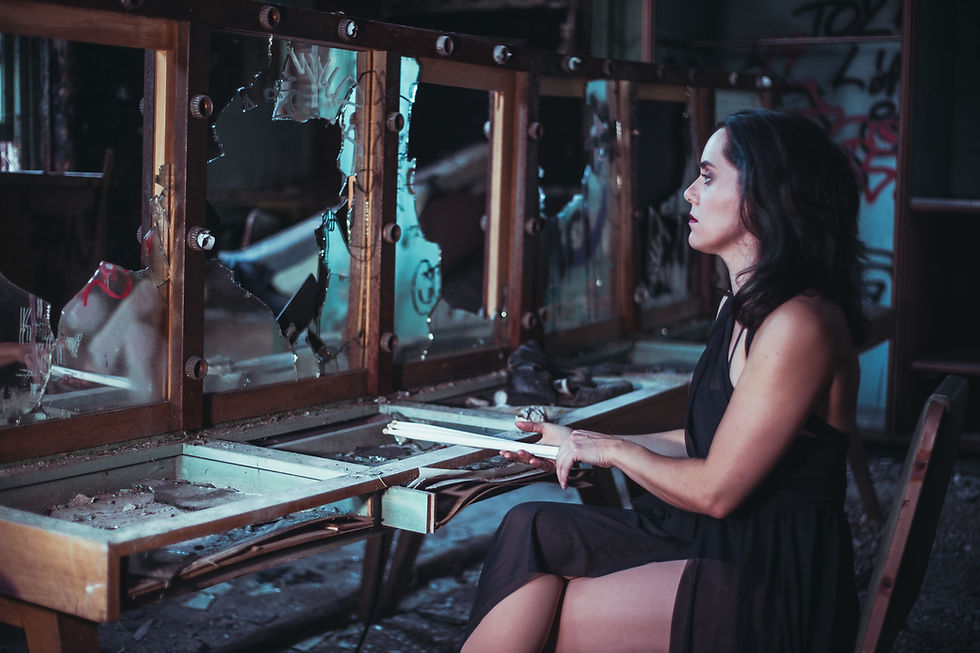 A female drummer juggler in a sheer, black dance dress sitting at a vandalized makeup table while holding drumsticks in a dilapidated dressing room.