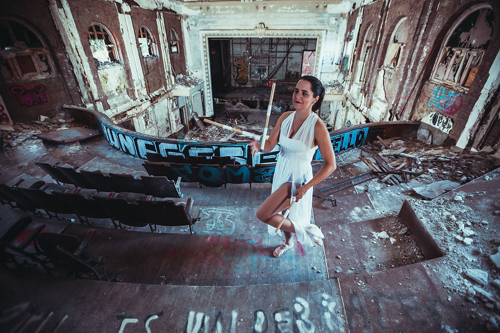 A female drummer juggler in a white dance dress juggling drumsticks in a dilapidated, graffiti-ridden auditorium.