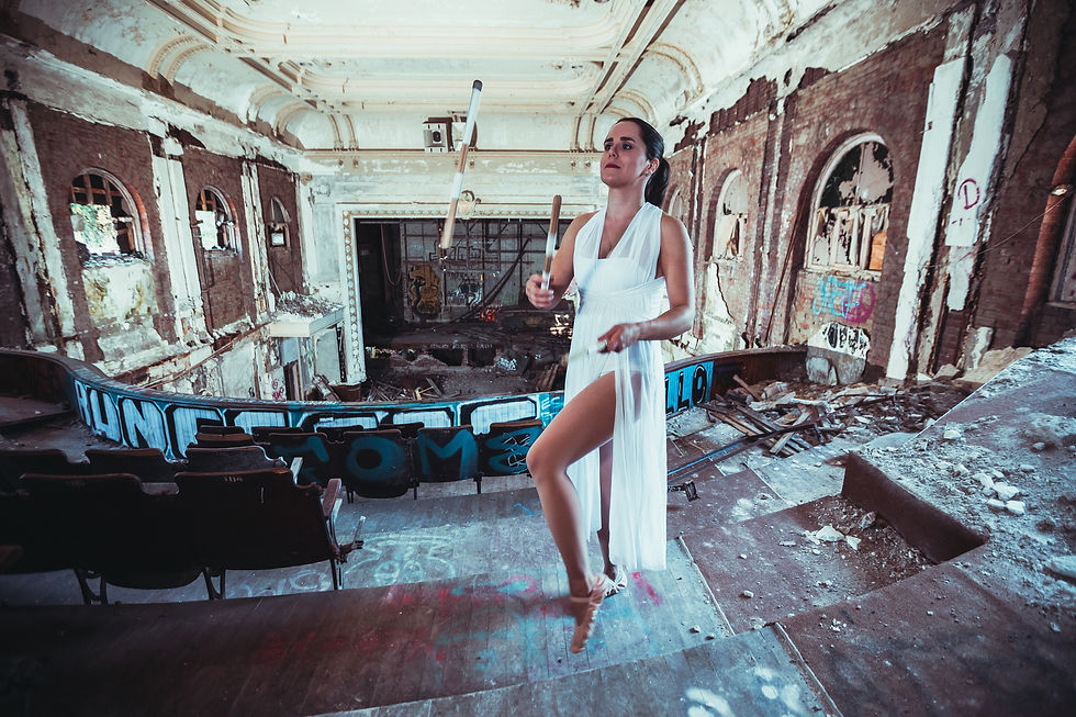 A female drummer juggler in a white dance dress juggling drumsticks in a dilapidated, graffiti-ridden auditorium.