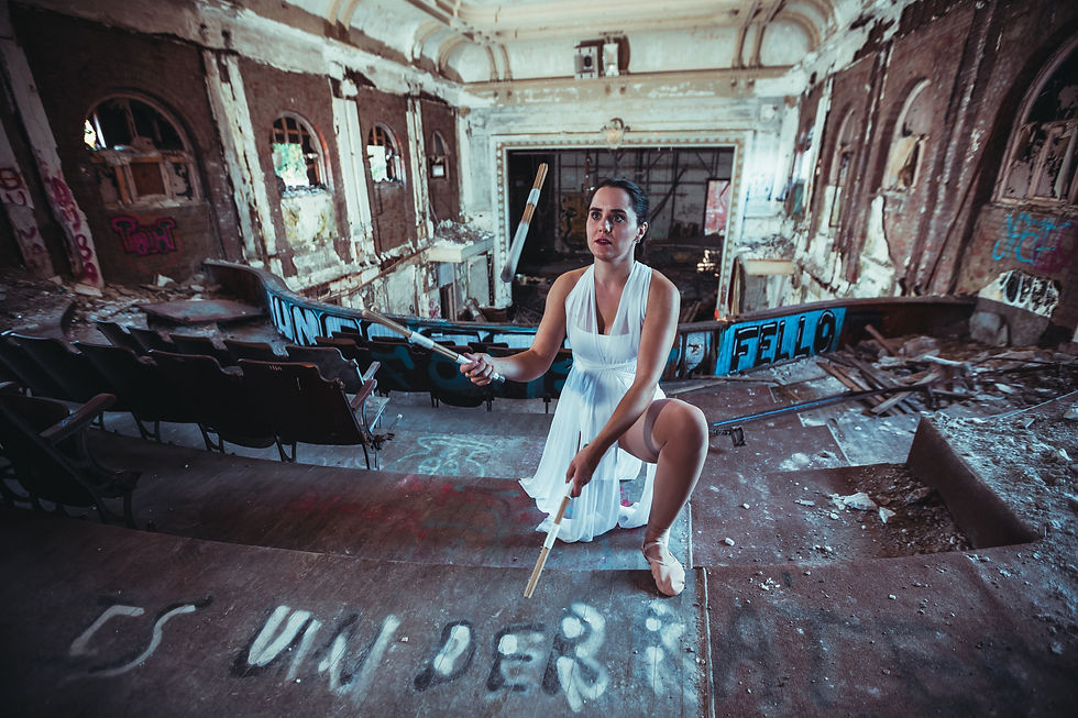 A female drummer juggler in a white dance dress juggling drumsticks in a dilapidated, graffiti-ridden auditorium.