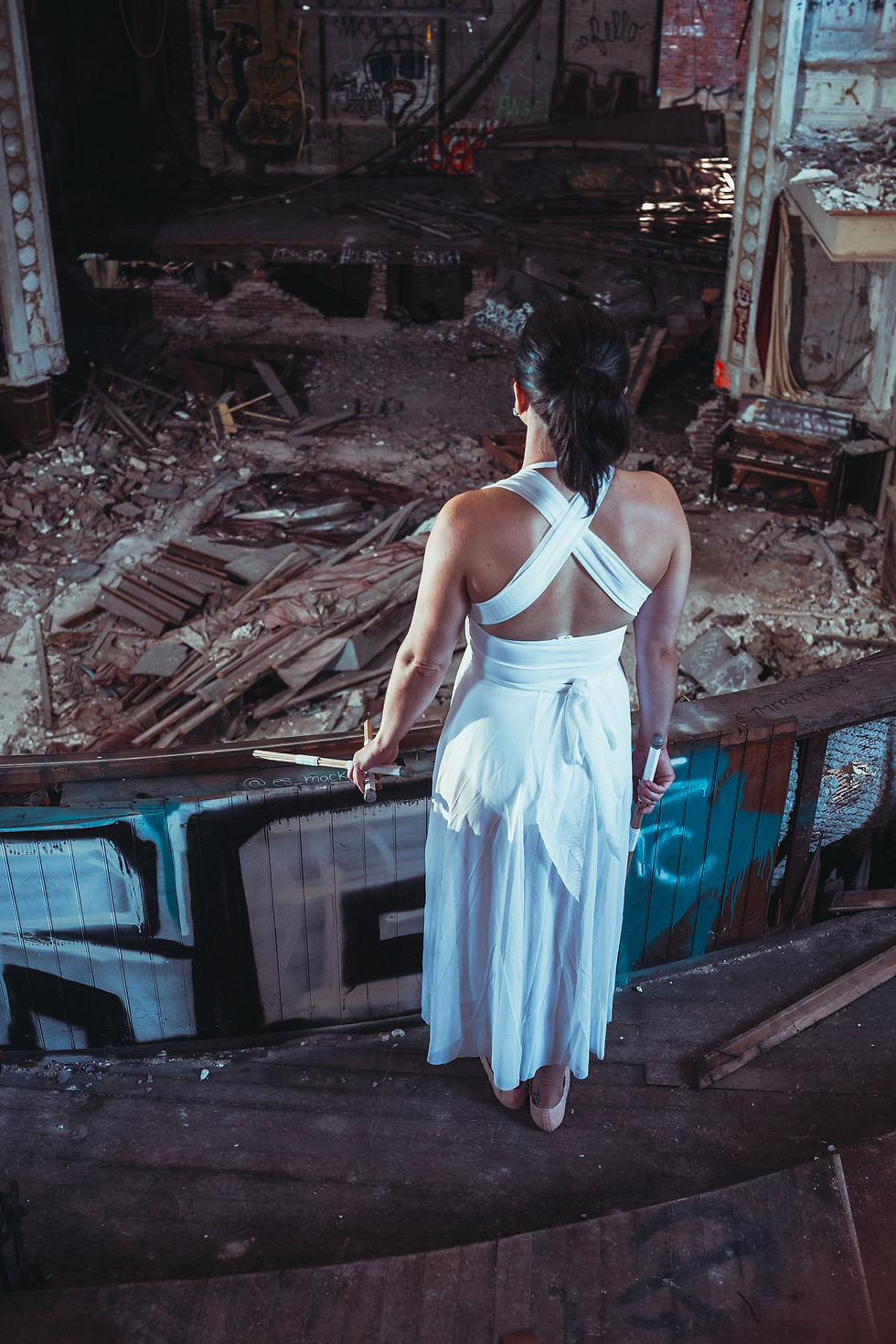A female drummer juggler in a sheer, white dance dress holding drumsticks while looking down into a dilapidated, graffiti-ridden auditorium.